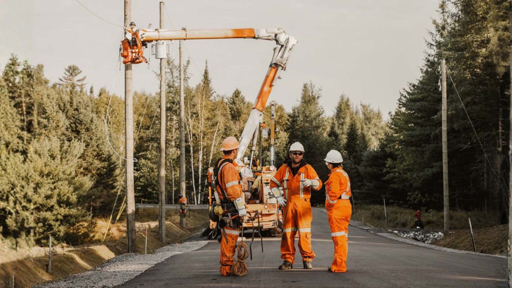 Trois travailleurs de Lanauco en uniforme orange discutent sur un chantier routier près d'une nacelle élévatrice, illustrant la marque employeur et la culture d'équipe sur le terrain.
