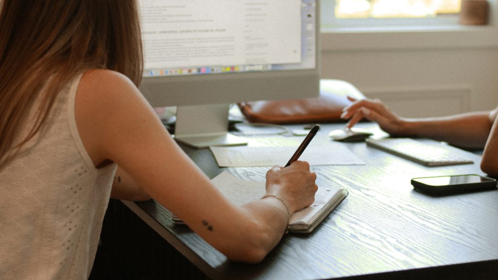 Bianka, stratège et fondatrice de l'agence Effet B à Saint-Sauveur, de dos à son bureau, prenant des notes manuscrites lors de l'analyse trimestrielle des données et de la stratégie marketing d'un client sur un ordinateur de bureau.